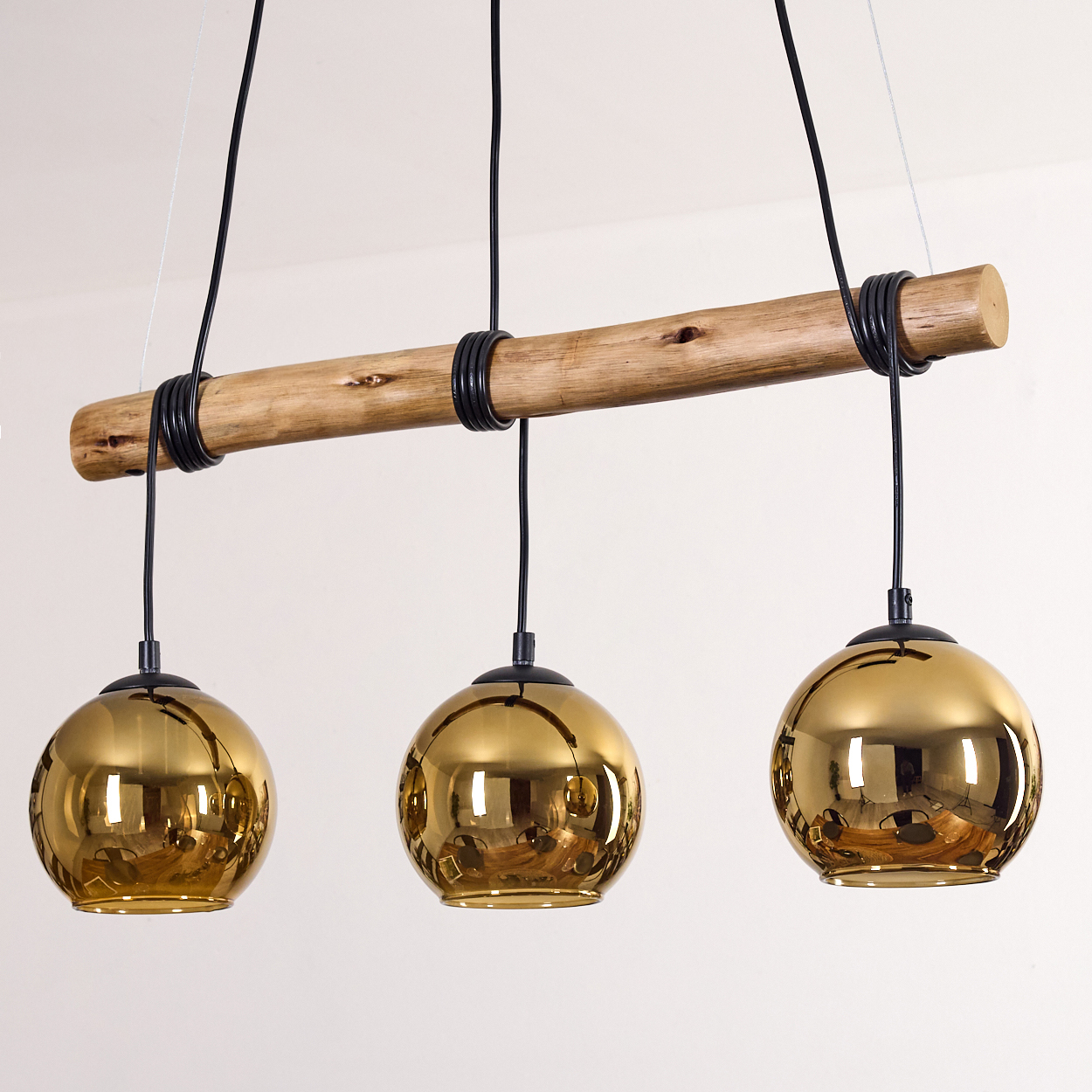 Eine moderne Deckenleuchte mit drei goldenen Kugellampen, die an einem Holzbalken hängen. Schwarze Drähte und weißer Hintergrund.