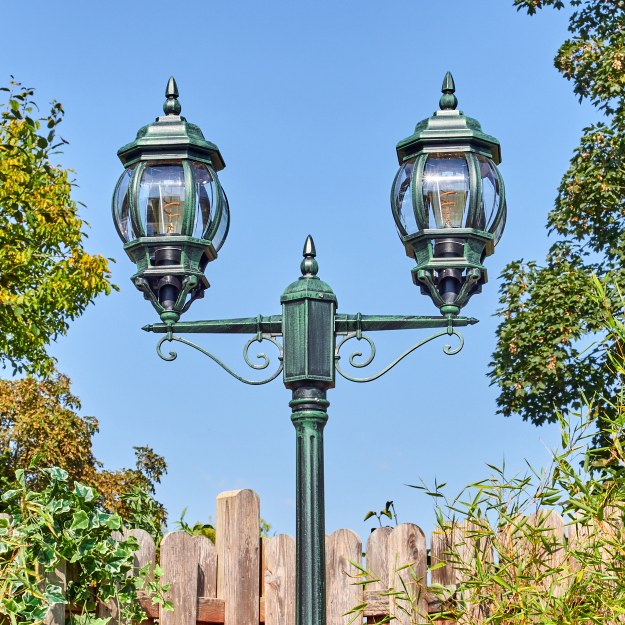 Eine grüne Straßenlaterne mit zwei Lampen. Der Hintergrund umfasst Bäume, einen Holzzaun und einen blauen Himmel.