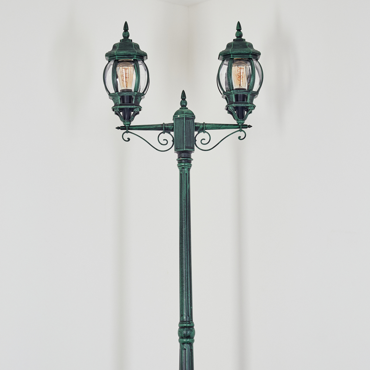 Eine grüne Straßenlaterne mit zwei Lichtern, jeweils mit einer Glühbirne. Die Lampe steht vor einer weißen Wand.