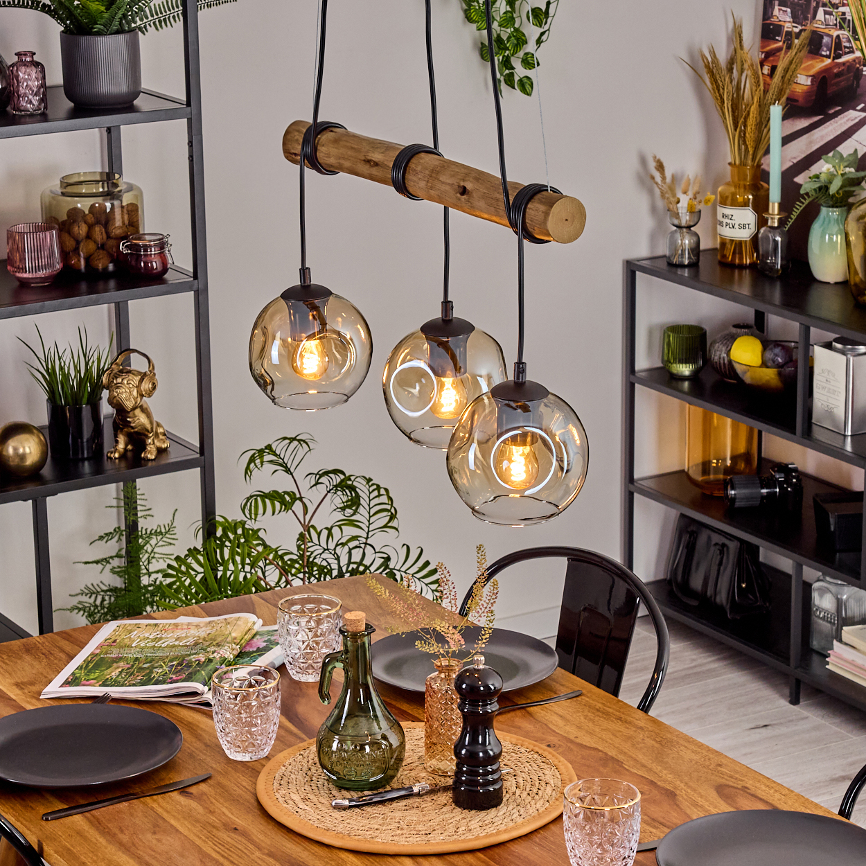 Esszimmer-Setup. Ein Holztisch ist mit Tellern, Gläsern und einer grünen Flasche gedeckt. Darüber hängen drei kugelförmige Lichter von einem Holzbalken.