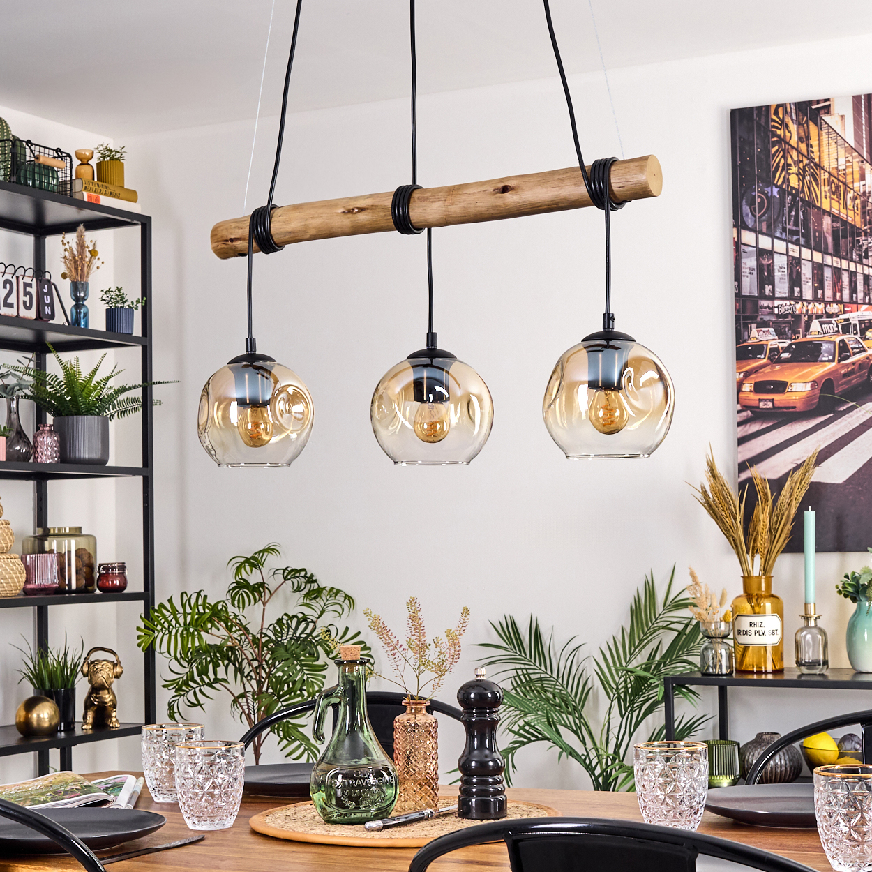 Drei-Leuchten-Pendelleuchte über einem Holztisch, der zum Abendessen gedeckt ist. Pflanzen, Gläser und ein Foto von New York City sind im Hintergrund.