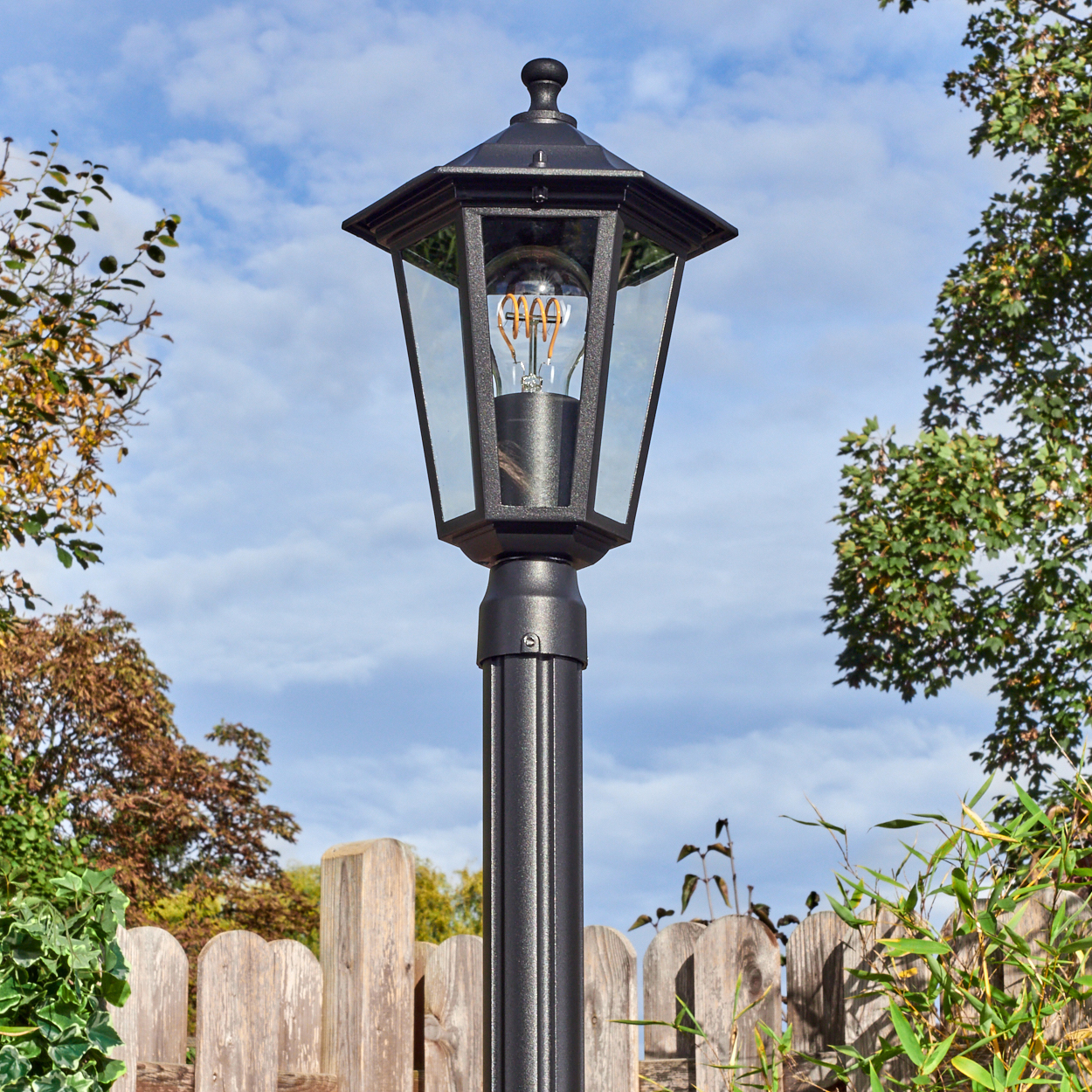 Eine schwarze Außenlampe mit sichtbarer Glühbirne steht vor einem Holzzaun. Bäume und blauer Himmel mit Wolken im Hintergrund.