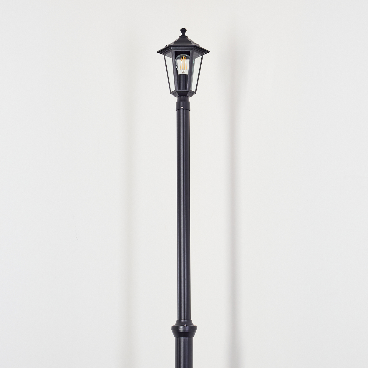 Eine schwarze Straßenlaterne steht vor einer weißen Wand. Die Lampe leuchtet. An der Wand ist ein Schatten zu sehen.