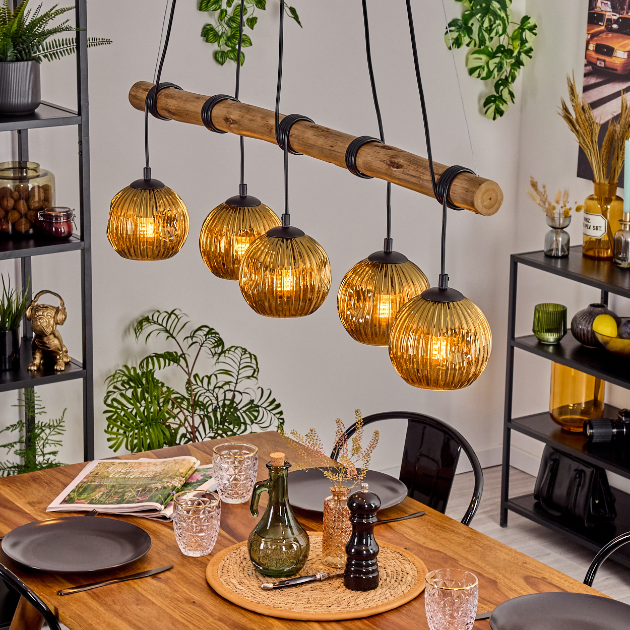 Ein Esstisch mit Geschirr, Gläsern und einem Mittelstück. Darüber hält ein Holzbalken fünf goldene Kugelleuchten.