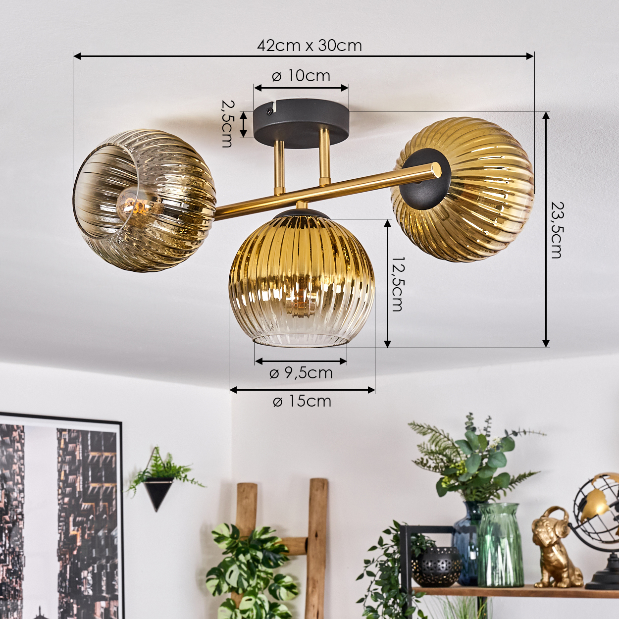 Moderne Deckenleuchte mit drei goldenen Glasschirmen. Maße sind angegeben. Pflanzen und Dekoration im Hintergrund.