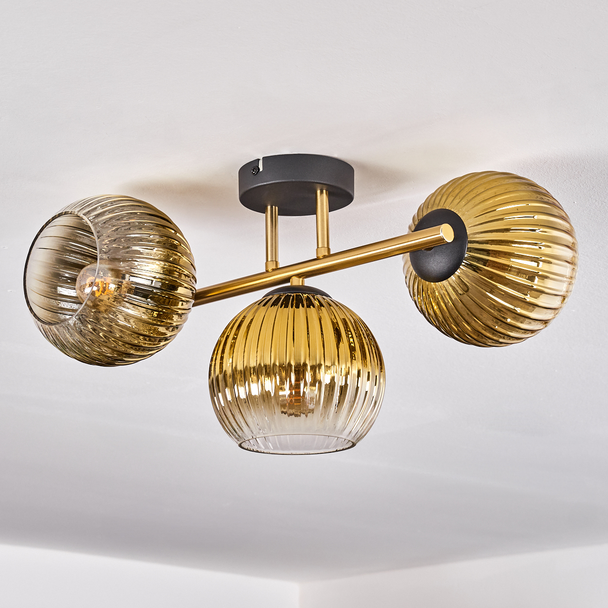 Eine moderne Deckenleuchte mit drei goldenen, gerippten Glaskugeln. Sie ist an einer weißen Decke mit schwarzem Sockel und goldenen Armen befestigt.