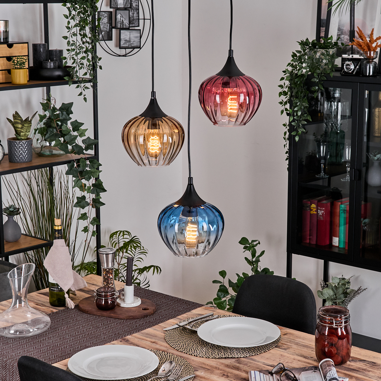 Esszimmer mit drei bunten Pendelleuchten. Tisch mit Tellern gedeckt. Regale mit Pflanzen, einem Schrank und einem Einmachglas.
