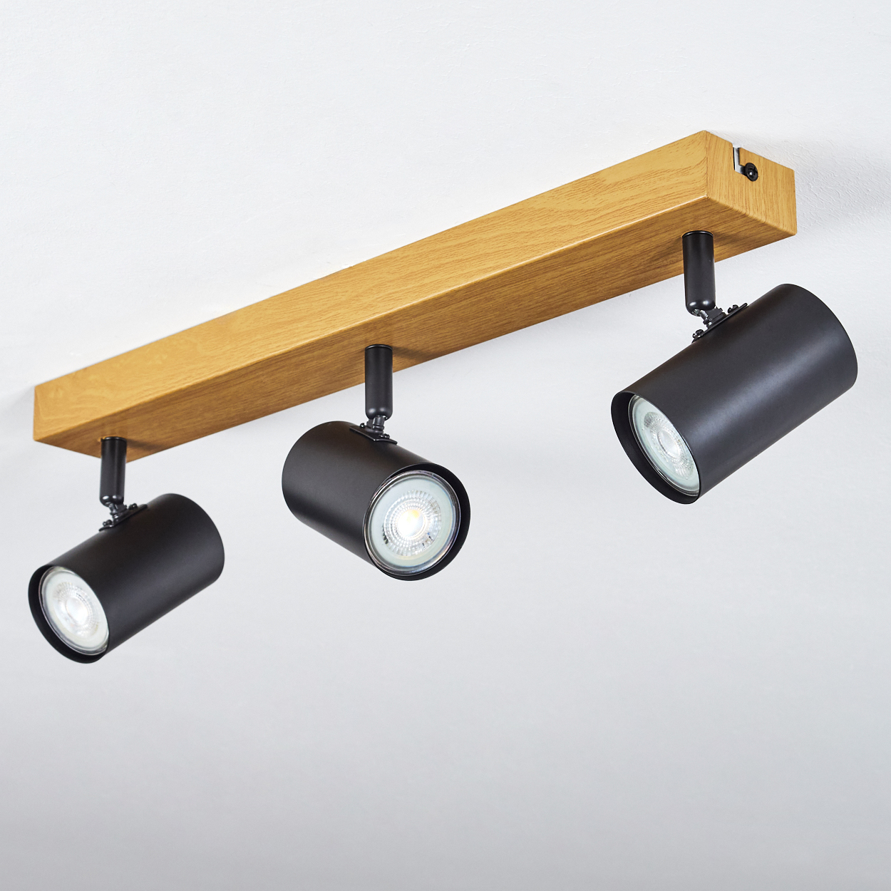 Drei schwarze Strahler an einem Holzbalken, an einer weißen Decke befestigt. Lichter sind an, beleuchten den Raum.