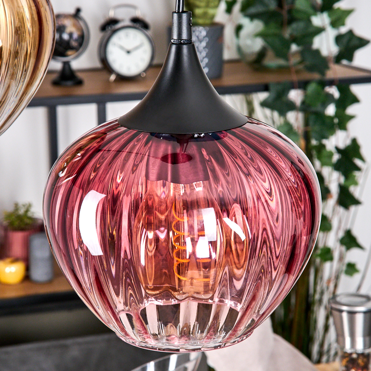Eine rosa Glas-Pendelleuchte mit geripptem Design, in der sich eine glühende Glühbirne befindet. Eine schwarze Halterung stützt die Lampe. Im Hintergrund befindet sich Dekoration.