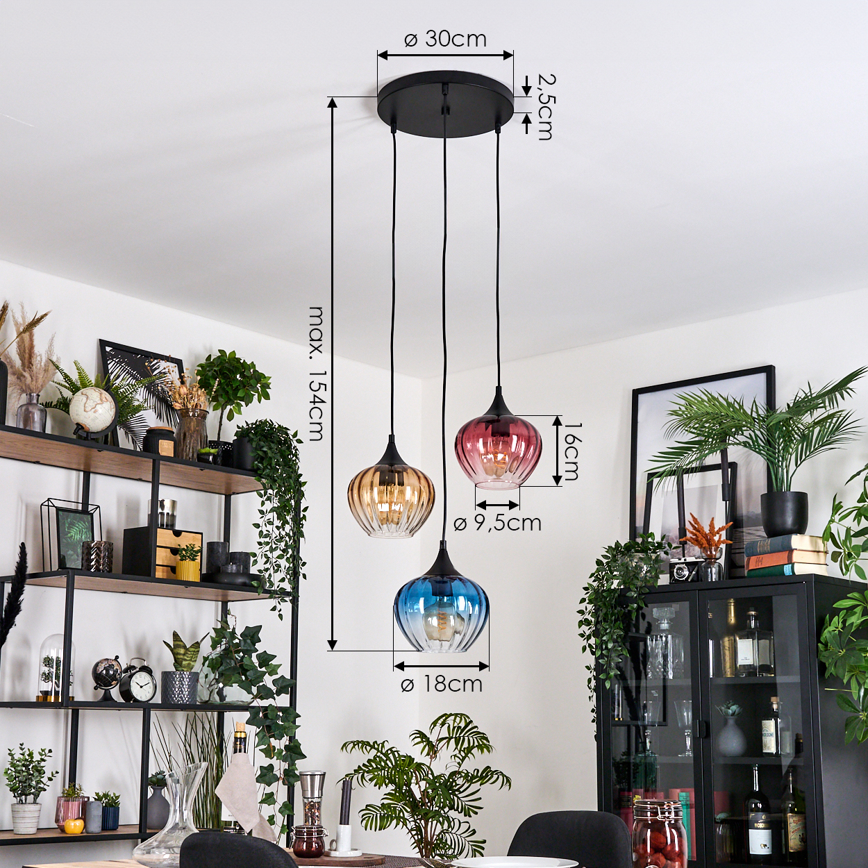 Drei farbige Pendelleuchten hängen von der Decke. Links ein Regal mit Deko und Pflanzen. Rechts ein Schrank mit Pflanzen.