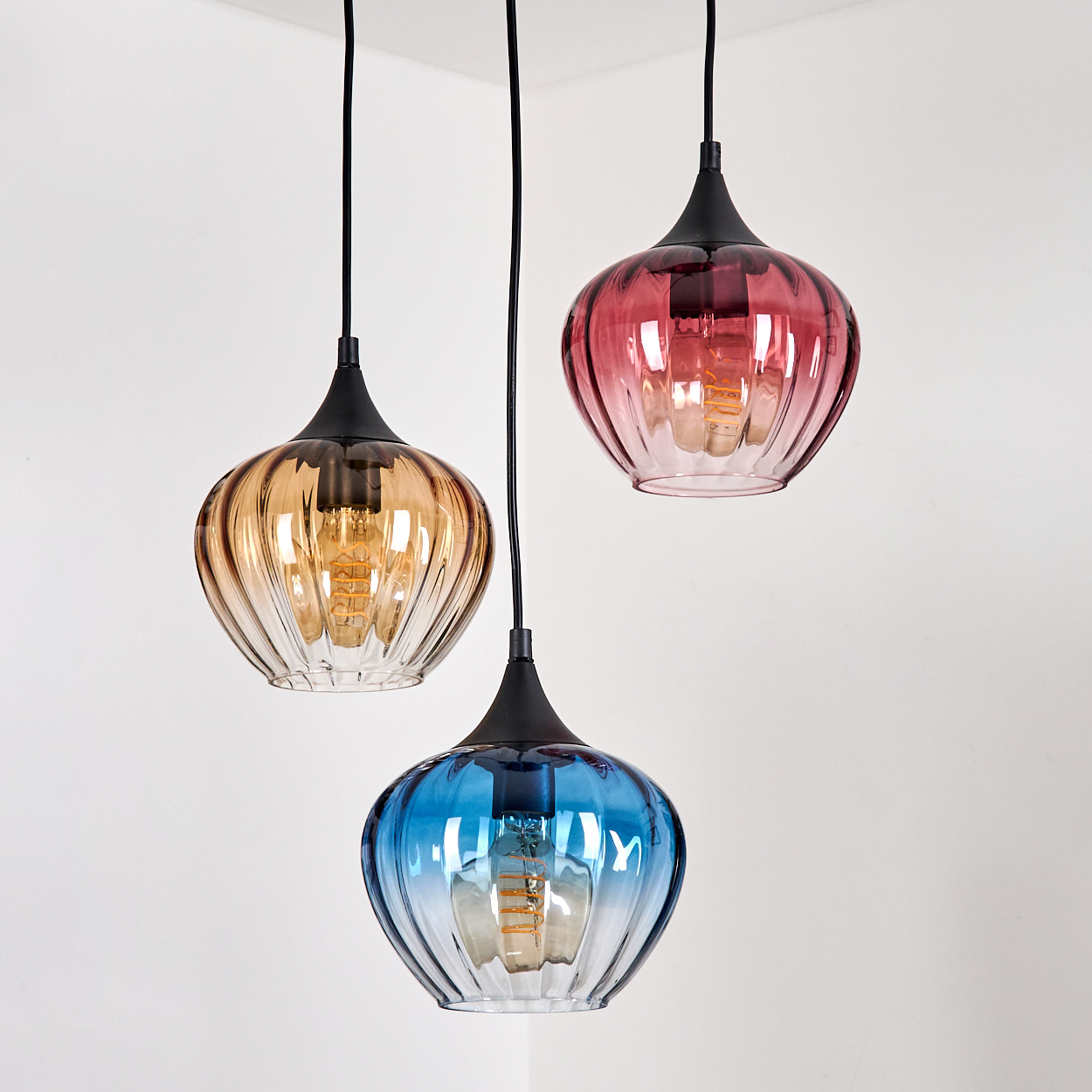 Drei farbige Glas-Pendelleuchten hängen vor einer weißen Wand. Die Lampen sind bernsteinfarben, rosa und blau, jede mit einer sichtbaren Glühbirne.