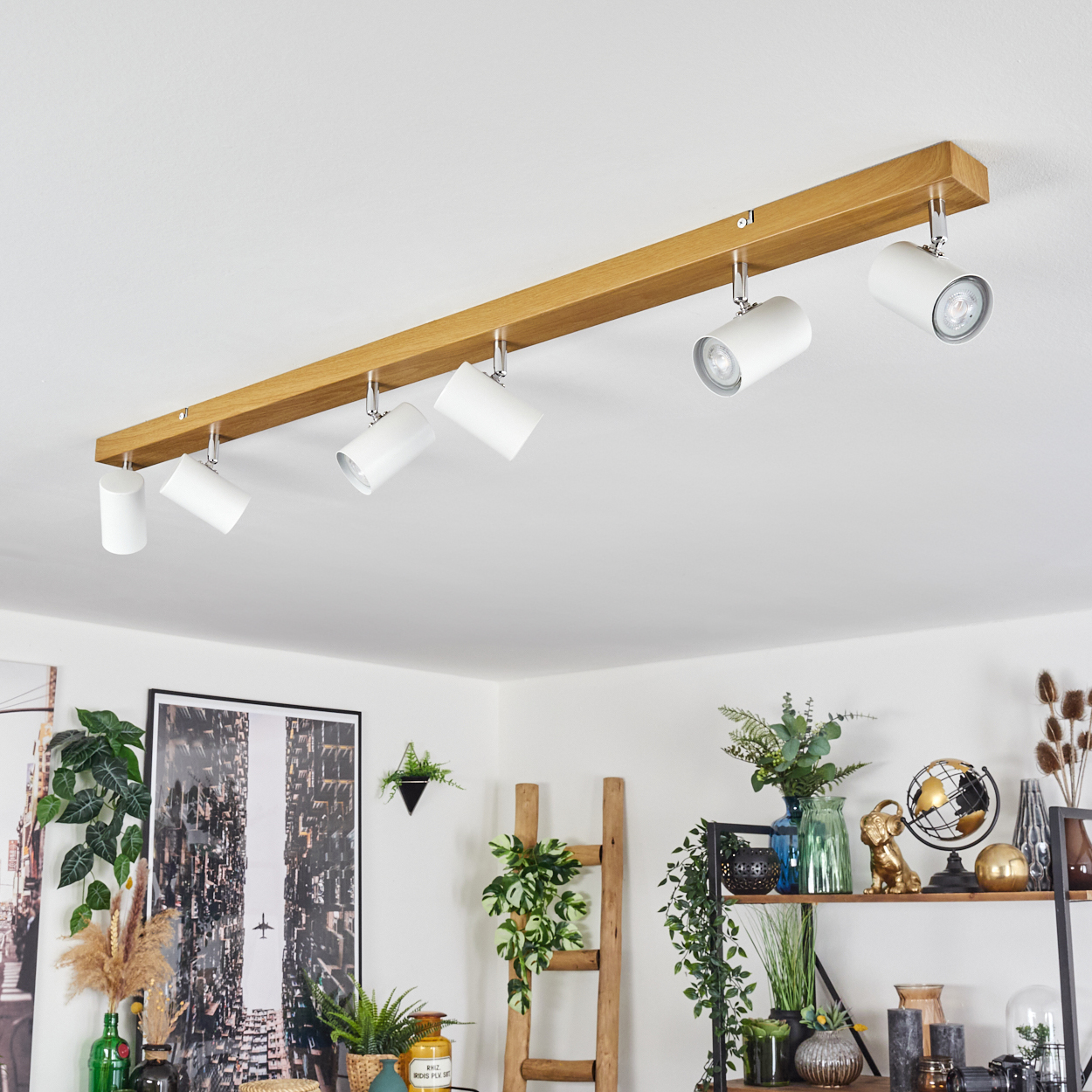 Ein Holzbalken mit sechs weißen Strahlern an der Decke. Darunter ein Raum mit Pflanzen, einer Leiter, Regalen und Kunst dekoriert.