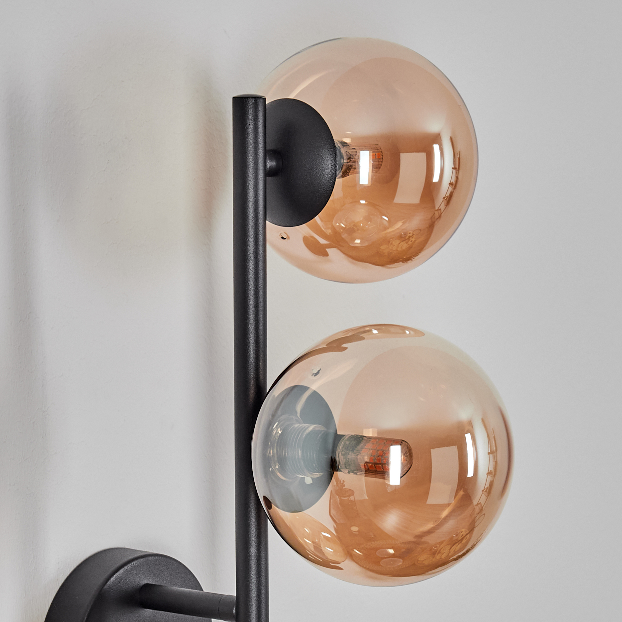 Eine moderne Wandlampe mit zwei kugelförmigen, bernsteinfarbenen Glasschirmen. Die Lampe hat einen schwarzen Metallrahmen und ist an einer weißen Wand befestigt.
