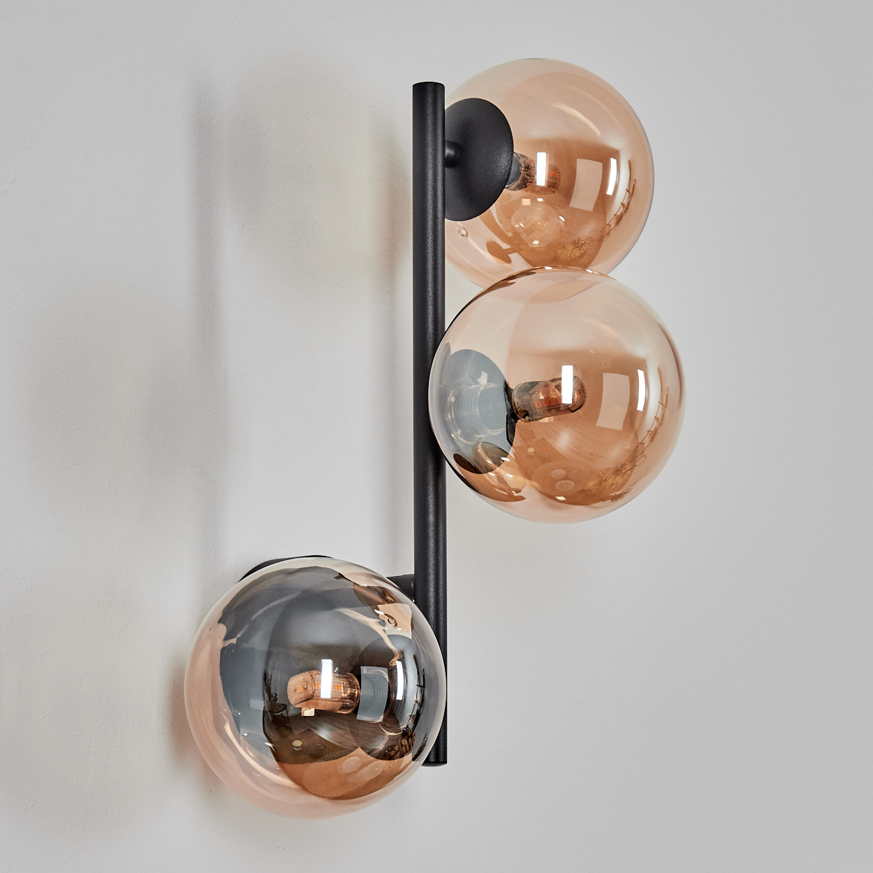 Eine moderne Wandlampe mit drei kugelförmigen, bernsteinfarbenen Glasschirmen. Sie hat einen schwarzen Metallrahmen. Der Hintergrund ist eine weiße Wand.