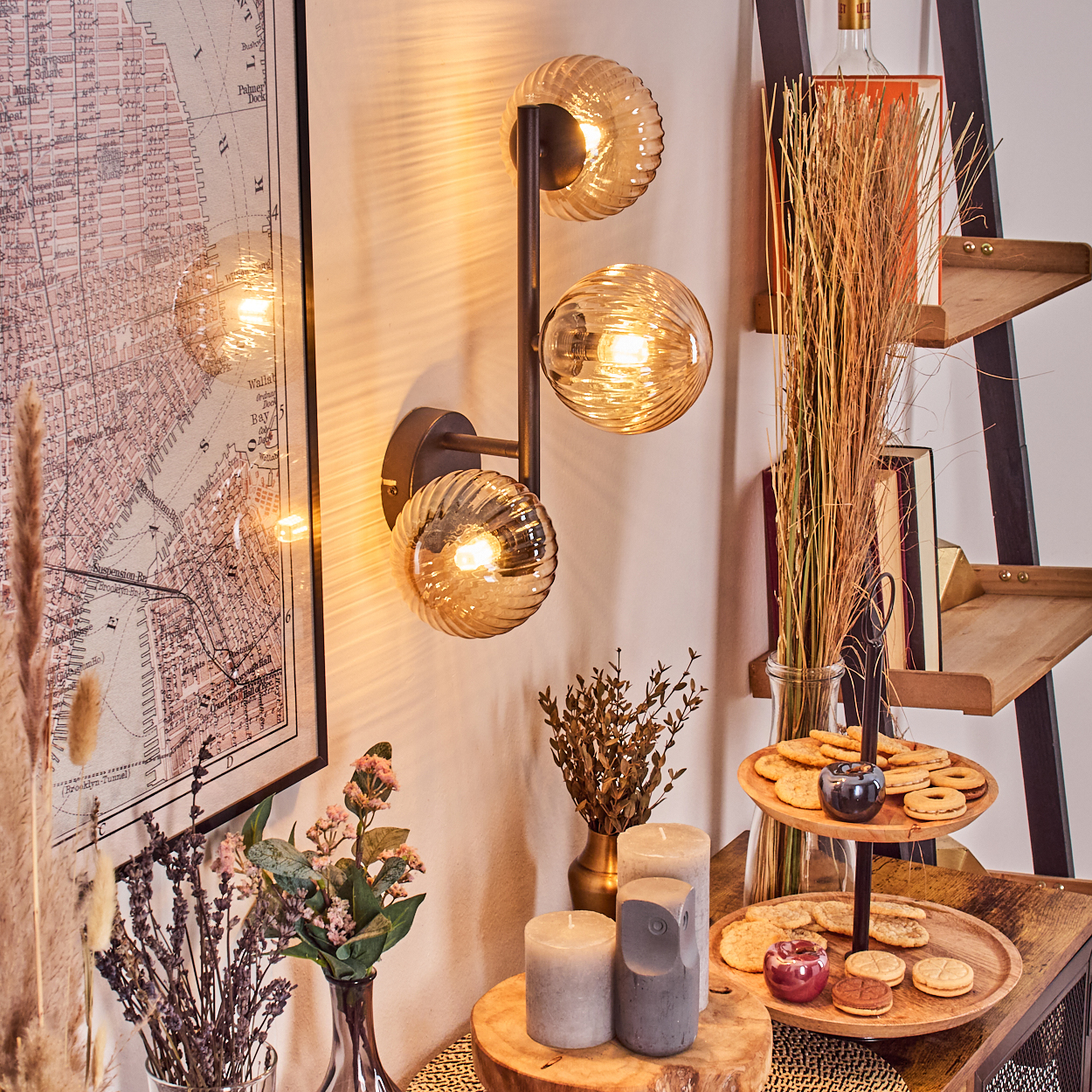 Eine dekorative Wandlampe mit drei Glaskugeln. Daneben ein Holzregal mit Büchern und Keksen. Eine Karte und Blumen sind ebenfalls zu sehen.