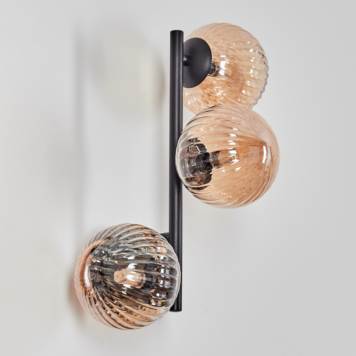 Dreifache Leuchte mit bernsteinfarbenen Glaskugeln an einem schwarzen Metallrahmen, an einer weißen Wand befestigt. Die Kugeln haben ein geripptes Design.
