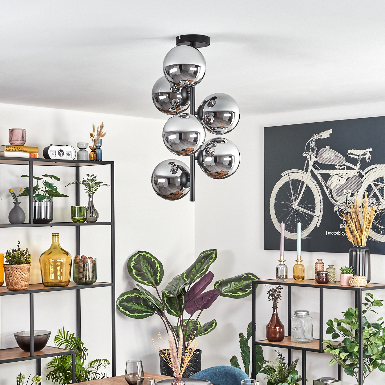 Ein modernes Zimmer mit einem Kronleuchter aus Chromkugeln, Pflanzen und einem Fahrradkunstwerk. Regale zeigen Dekorationen und weitere Pflanzen.