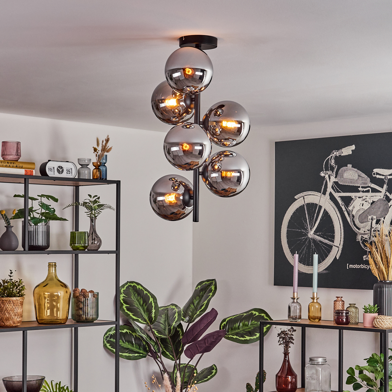Ein modernes Wohnzimmer mit einem Kronleuchter aus Chromkugeln. Regale mit Pflanzen und Dekoration. Ein Motorrad-Kunstwerk an der Wand.