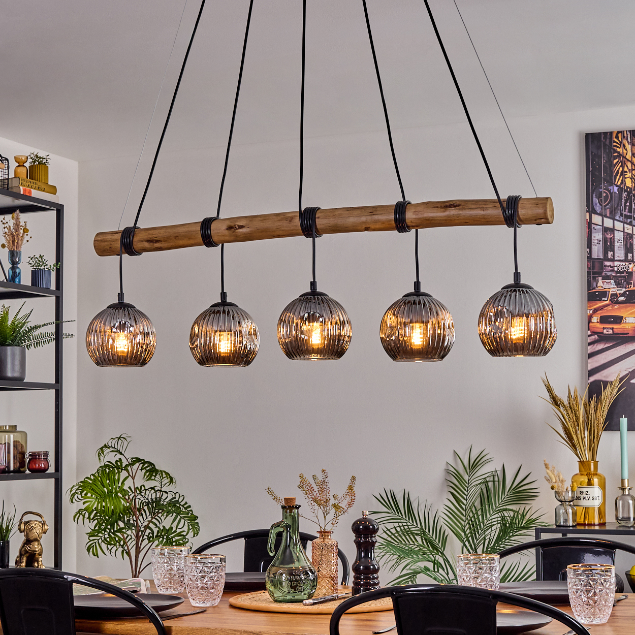 Ein Esszimmer mit einem Holzbalken und fünf Hängelampen. Ein Tisch ist gedeckt mit Gläsern und Pflanzen.