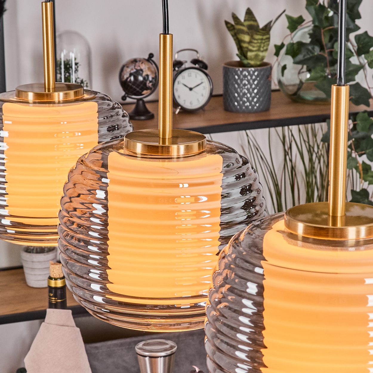 Drei Pendelleuchten mit Glasschirmen. Hintergrund mit Regal, Uhr, Globus, Pflanzen. Warme Beleuchtung.