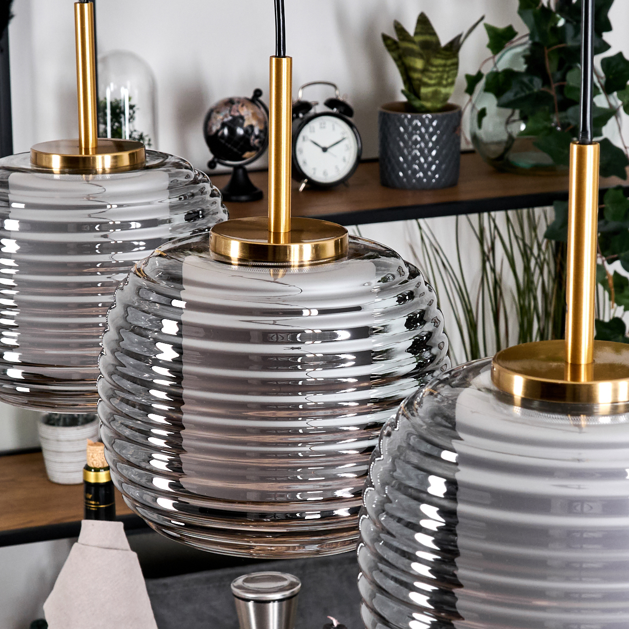 Drei Glaskugel-Pendelleuchten hängen. Im Hintergrund ein Regal mit Pflanzen, Uhr und Globus. Lampen haben goldene Armaturen.