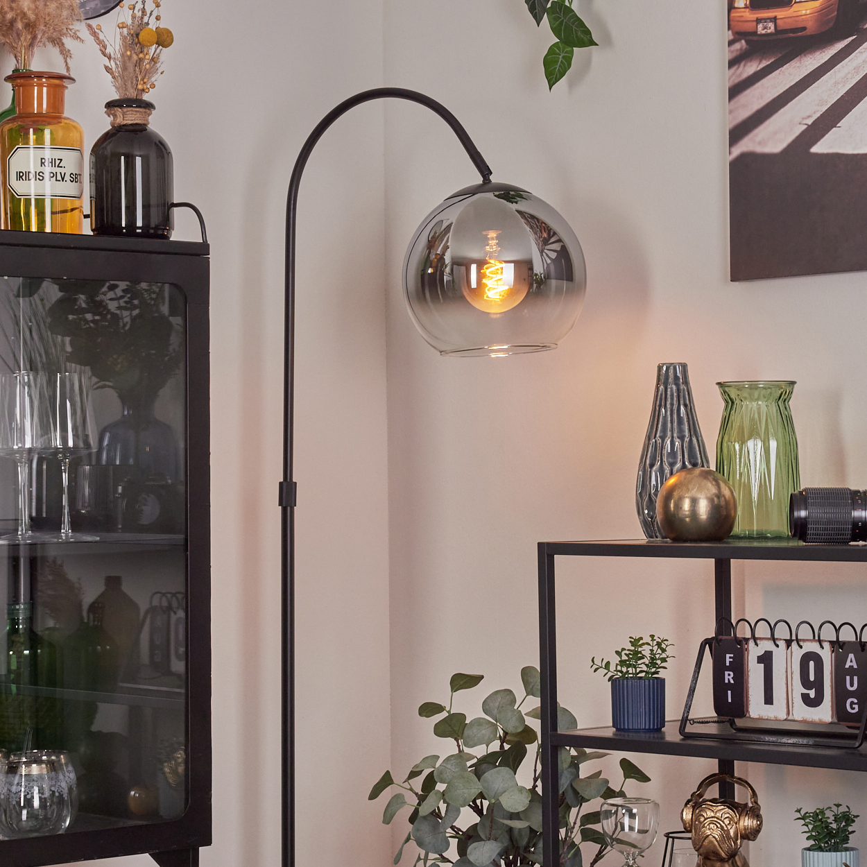 Ein modernes Wohnzimmer mit schwarzer Stehlampe, Glasvase und Regal. Ein Foto eines gelben Taxis hängt an der Wand.