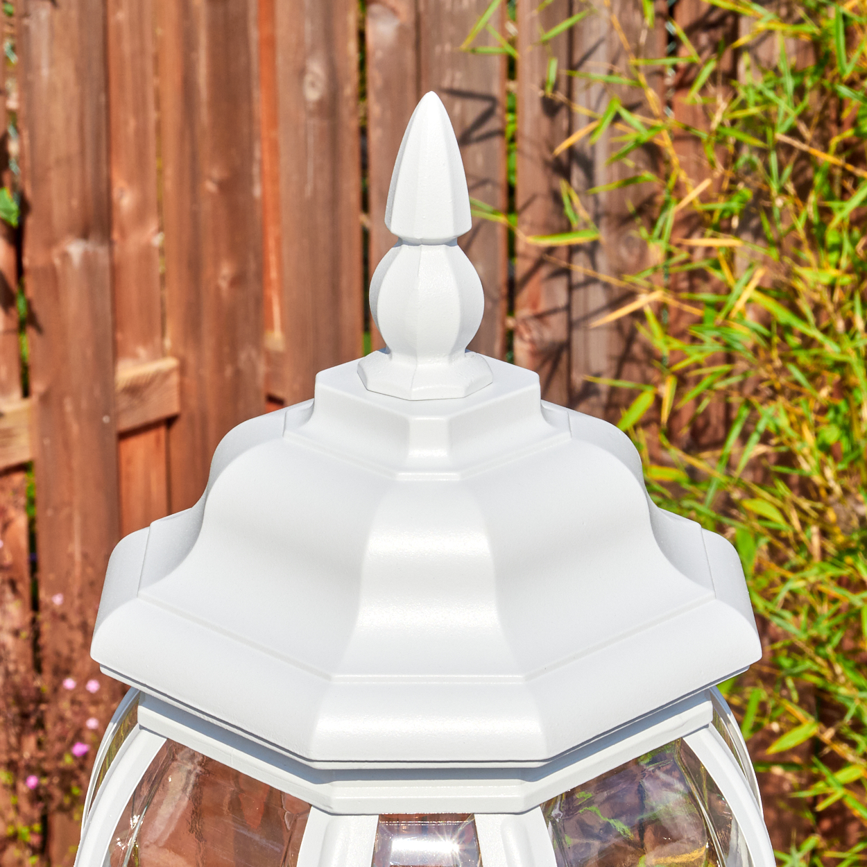 Nahaufnahme einer weißen Außenlampe mit spitzem Aufsatz. Im Hintergrund ein Holzzaun und grüne Pflanzen, beleuchtet von Sonnenlicht.