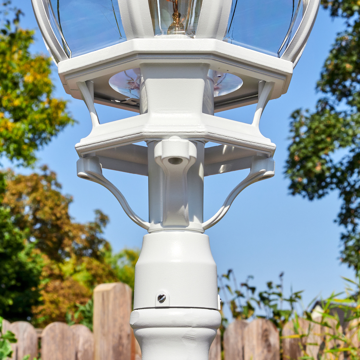 Eine weiße Außenlaterne mit Glaskugel. Der Hintergrund umfasst einen Holzzaun, Bäume und einen blauen Himmel.