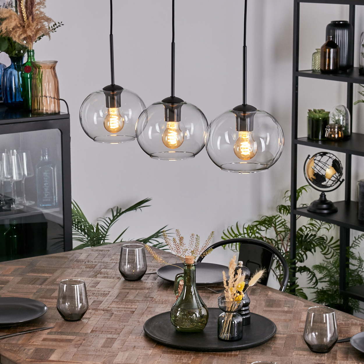 Esszimmer-Szene mit drei Hängelampen, einem Holztisch mit Tellern, Gläsern und Dekorationsartikeln.
