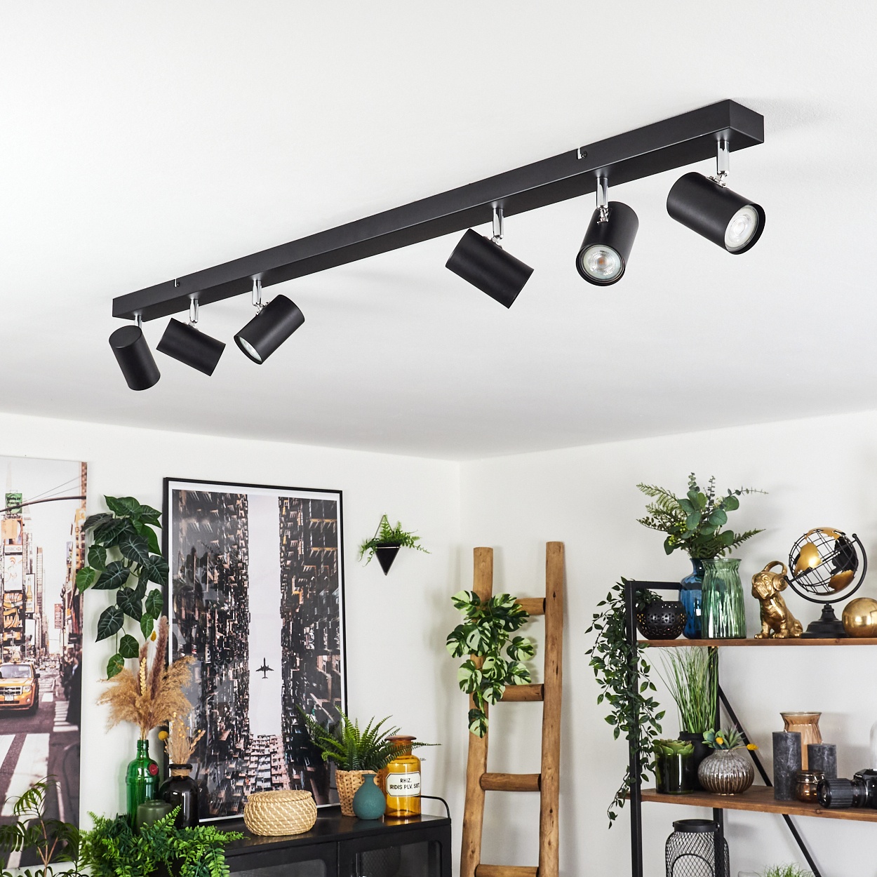 Schwarze Schienenbeleuchtung an einer weißen Decke. Darunter ist ein Raum mit Pflanzen, Kunstwerken, Regalen und einer Holzleiter eingerichtet.