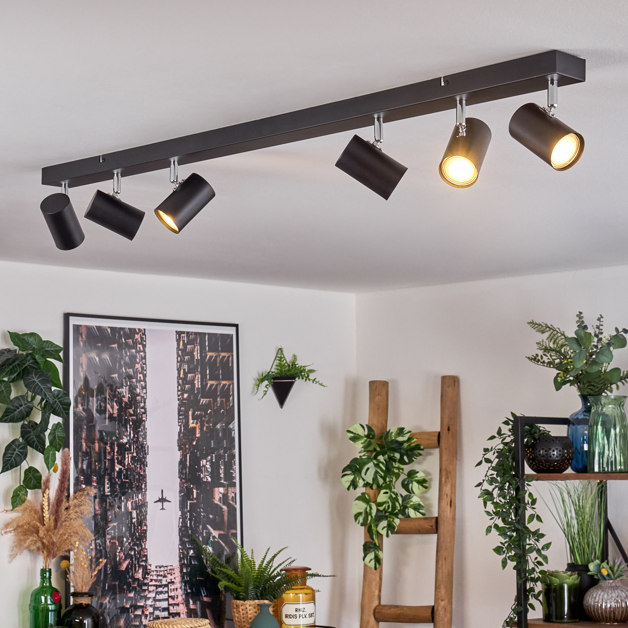 Eine Decke mit schwarzen Strahlern beleuchtet einen Raum. Ein Stadtbild hängt an der Wand. Pflanzen und eine Holzleiter sind im Raum.