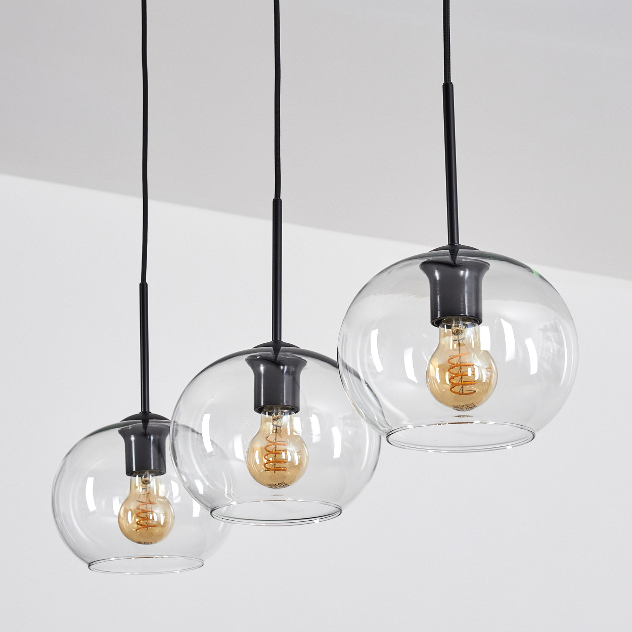 Drei Pendelleuchten mit Glaskugeln und sichtbaren Glühbirnen hängen von der Decke. Schwarze Armaturen und transparente Glasschirme.