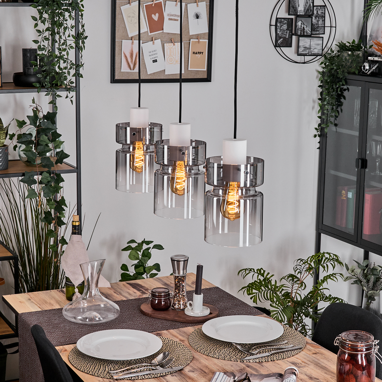 Esszimmer mit Holztisch, drei Hängelampen, Tellern, Pflanzen und einem schwarzen Schrank.