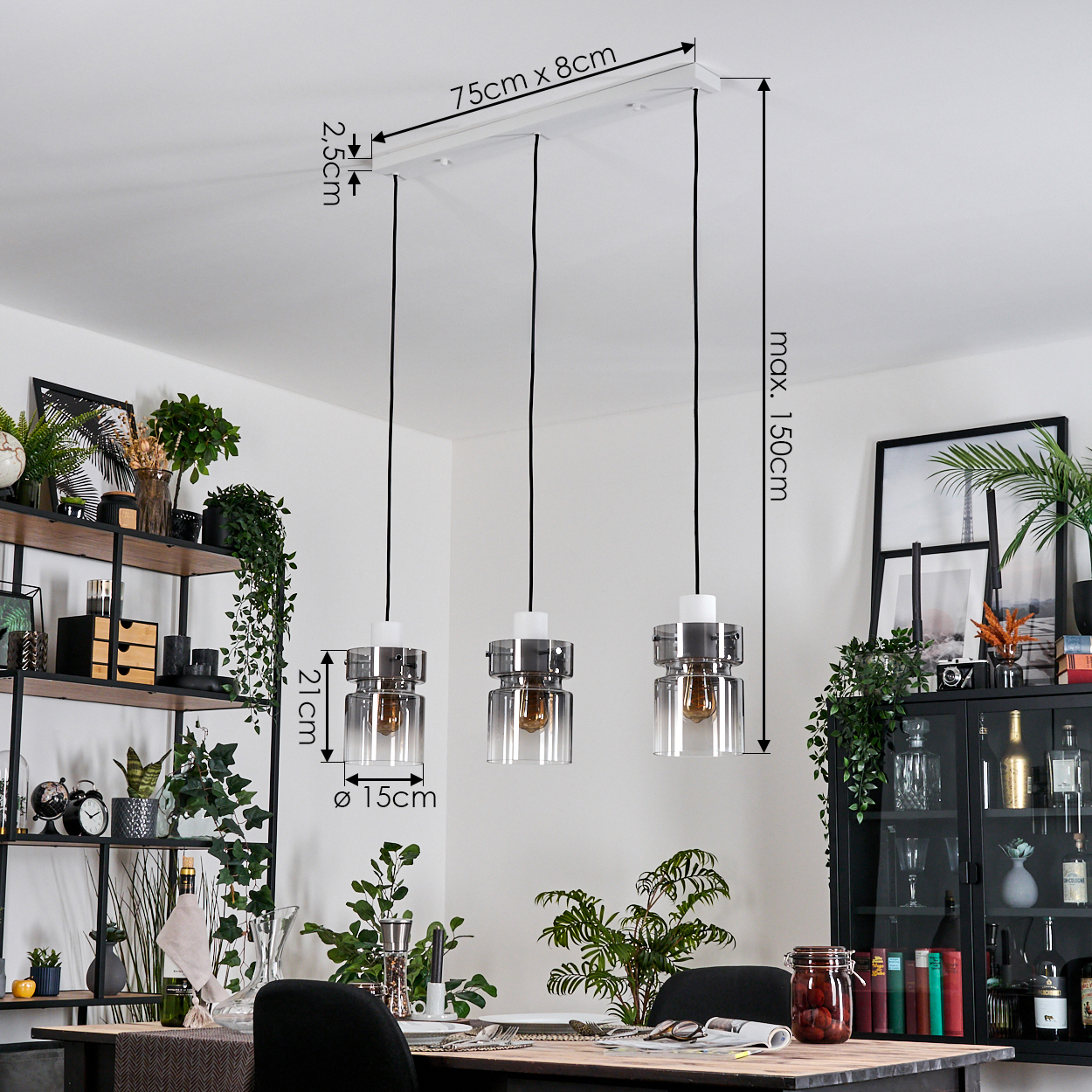 Drei Pendelleuchten hängen von der Decke. Ein Esstisch mit Pflanzen und Dekoration befindet sich im Vordergrund. Regale und ein Schrank sind sichtbar.