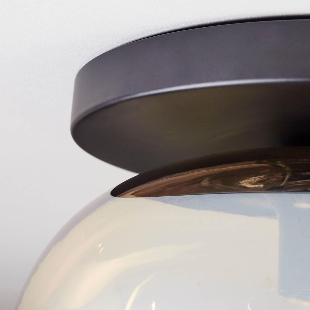 Nahaufnahme einer modernen Leuchte. Sie hat einen dunklen, kreisförmigen Metallsockel und eine Glaskuppel. Der Hintergrund ist eine weiße Decke.