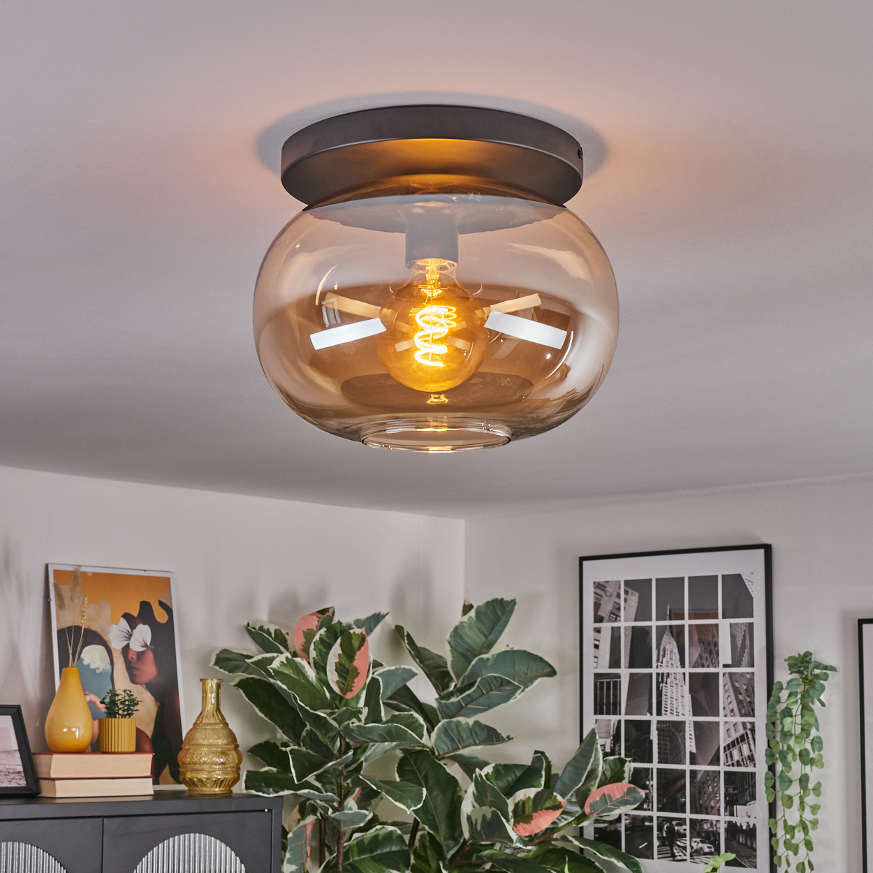 Eine moderne Deckenleuchte mit Glaskugel. Im Inneren befindet sich eine leuchtende Glühbirne, umgeben von einer weißen Decke. Pflanzen im Hintergrund.