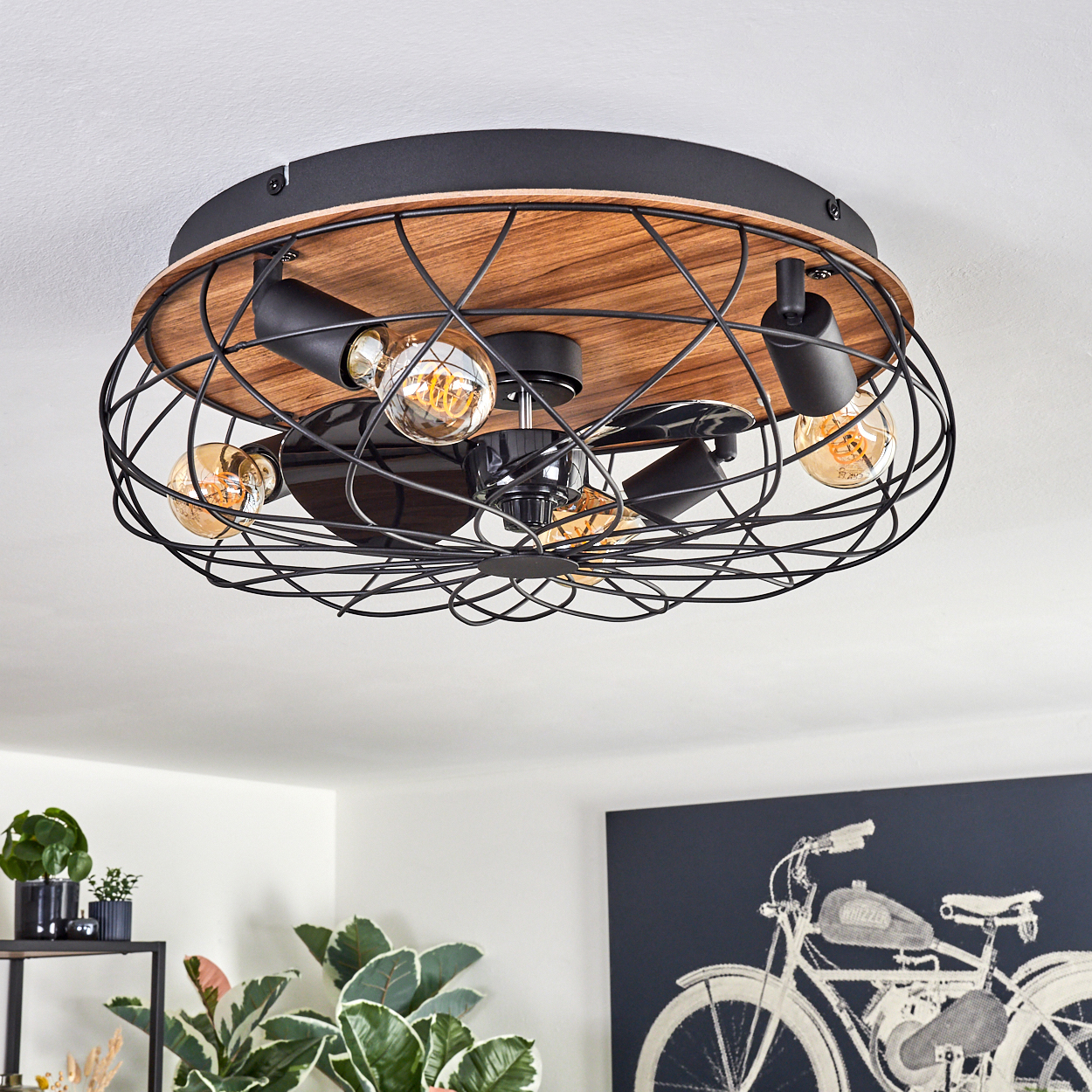 Eine Deckenleuchte mit Holzsockel, schwarzem Metallkäfig und freiliegenden Glühbirnen. Im Hintergrund sind Pflanzen und ein Motorradkunstwerk zu sehen.