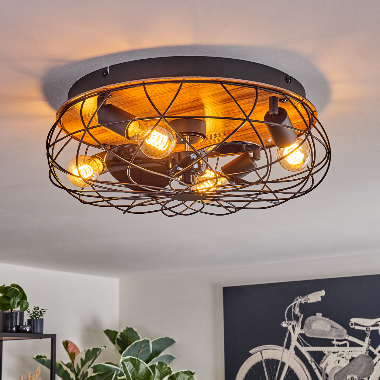 Eine Deckenleuchte mit freiliegenden Glühbirnen, umhüllt von einem Metallkäfig mit Holzsockel. Der Raum hat Pflanzen und Fahrradkunst.