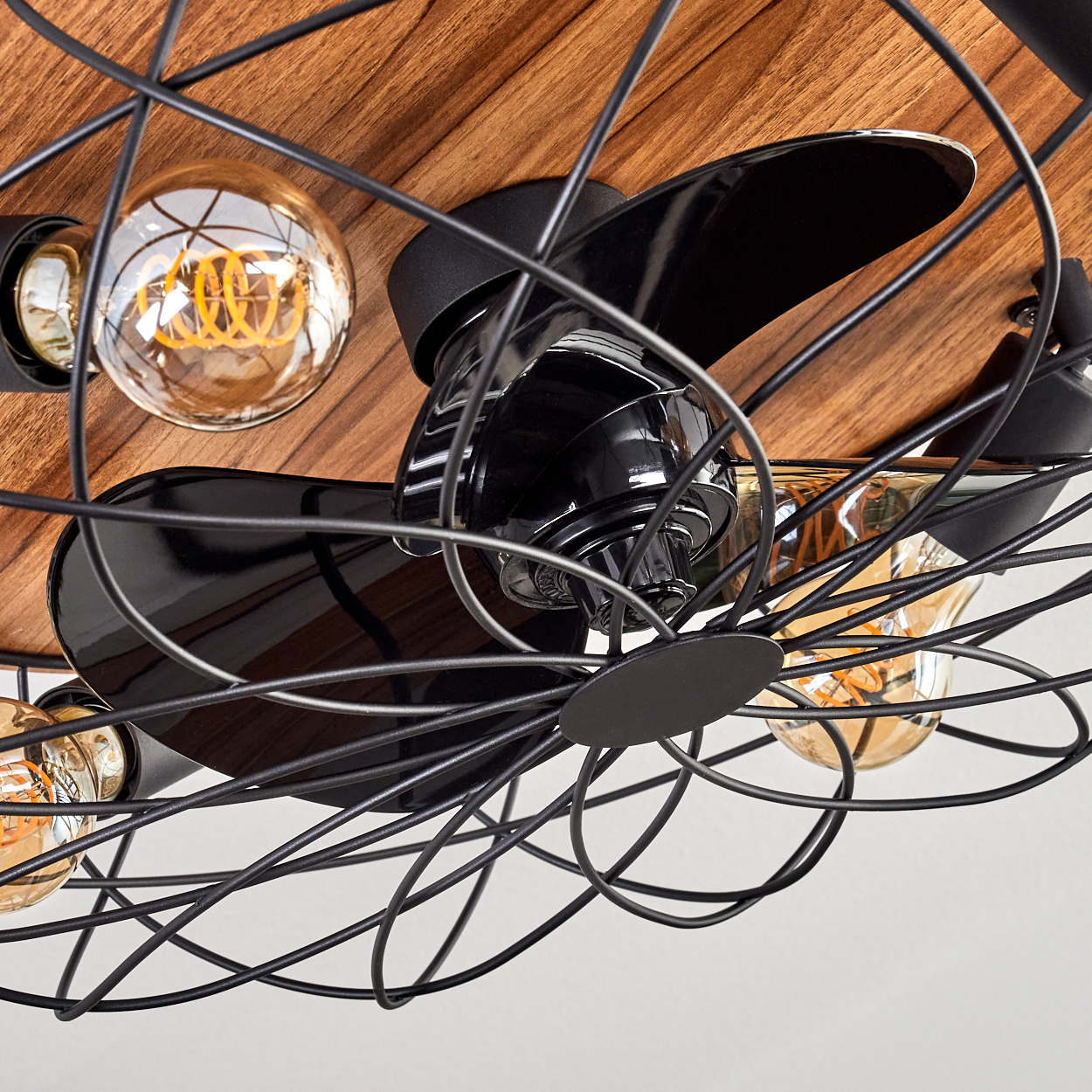 Nahaufnahme eines Deckenventilators mit schwarzen Flügeln und einem Drahtkäfig. Er hat Edison-Glühbirnen vor einem Holz Hintergrund.