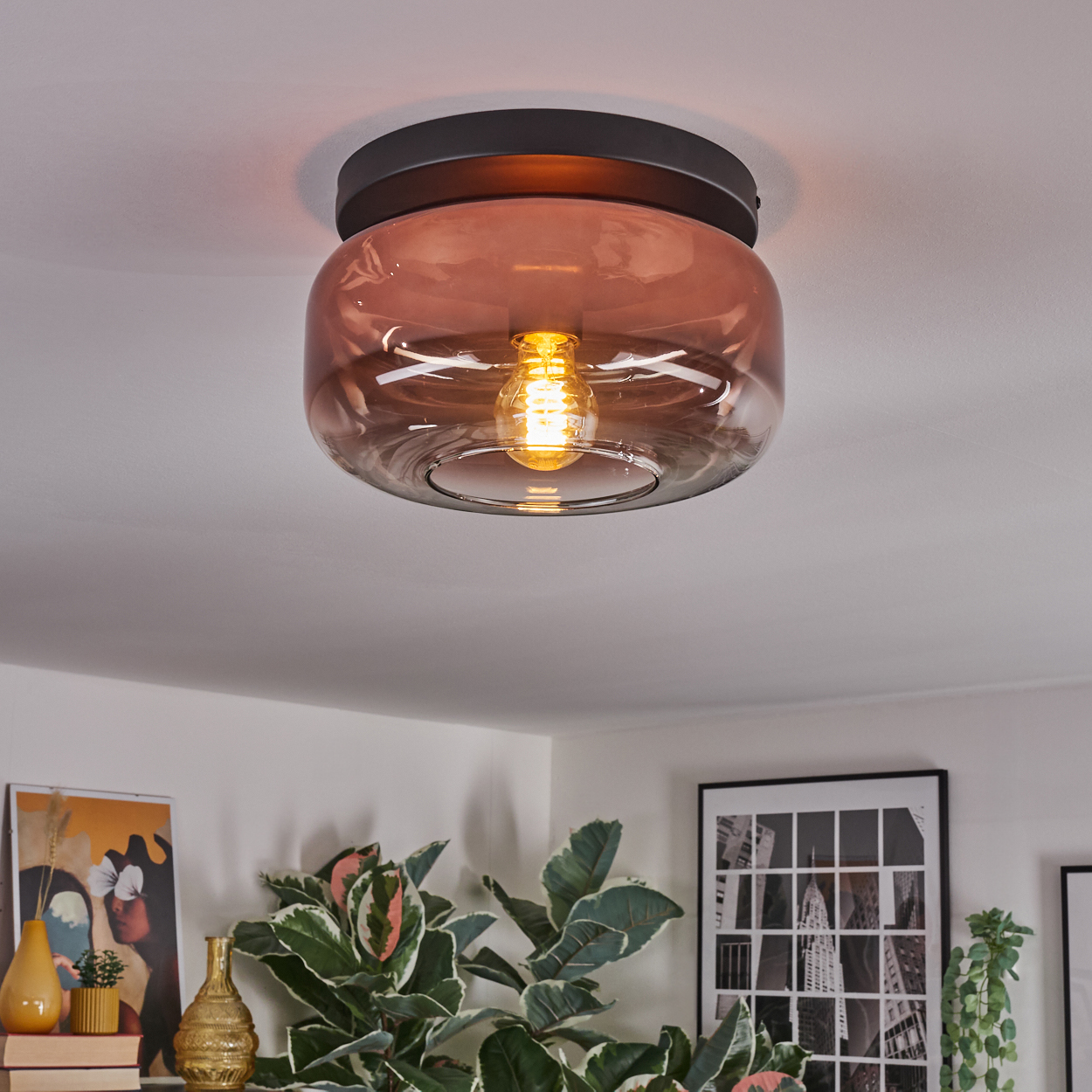 Eine moderne Deckenlampe mit getöntem Glasschirm. Sie beleuchtet einen Raum mit Kunstwerken und Pflanzen.