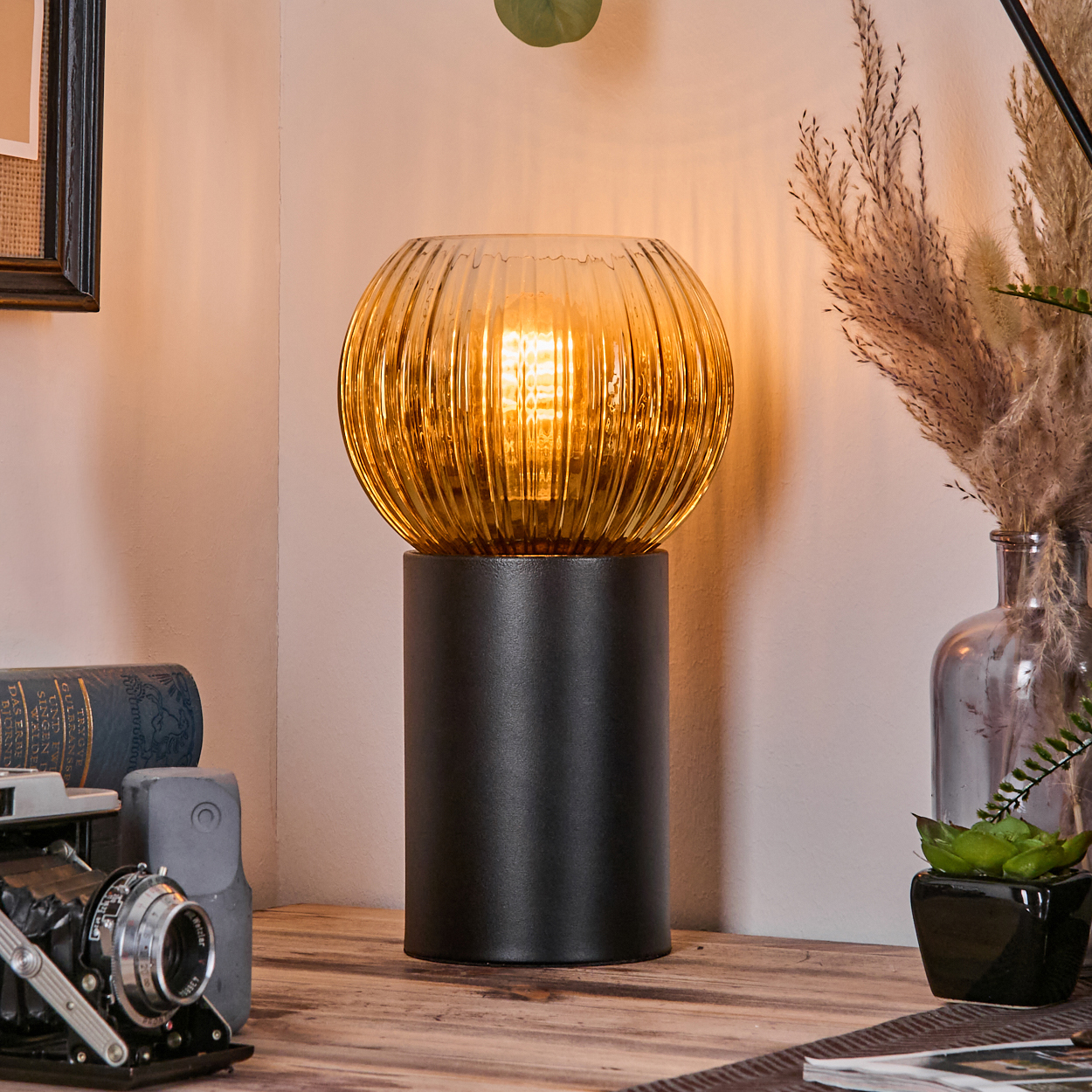 Eine leuchtende, bernsteinfarbene Glaslampe steht auf einem Holztisch. Eine Kamera, Bücher und eine Vase mit Trockenblumen sind sichtbar.