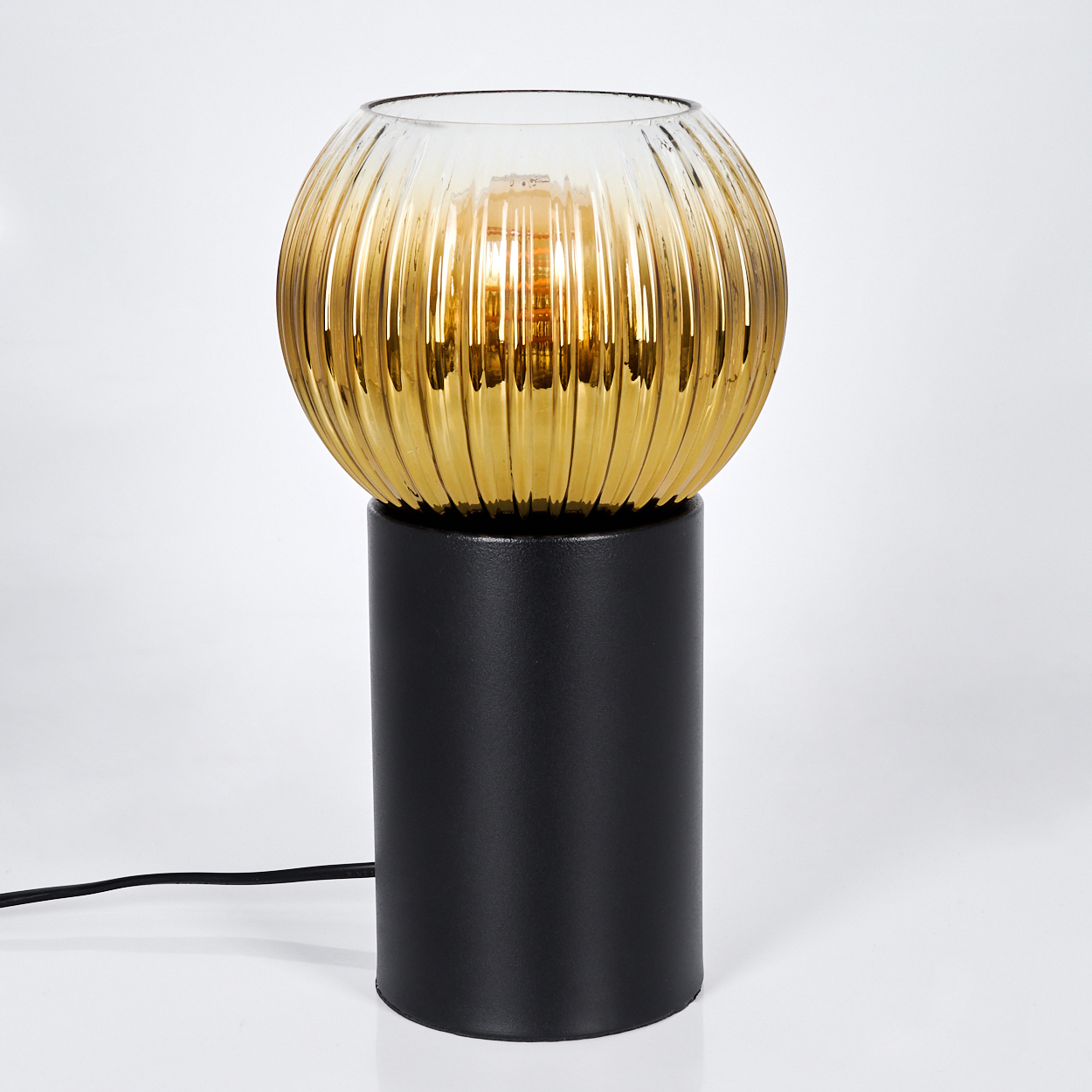 Moderne Lampe mit schwarzem zylindrischen Sockel und kugelförmigem, gerippten, bernsteinfarbenem Glasschirm. Die Glühbirne leuchtet innen. Weißer Hintergrund.