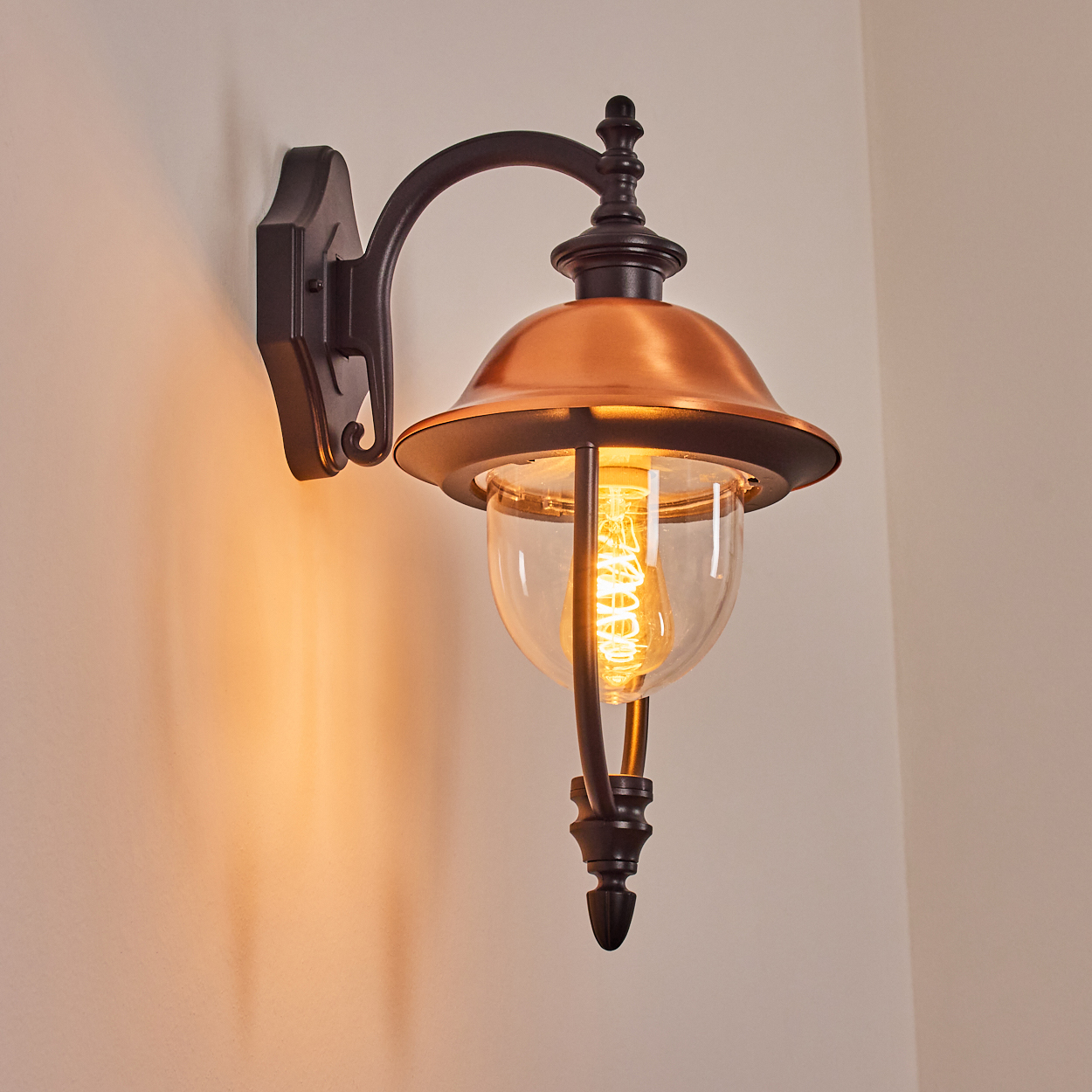 Eine dekorative Wandlampe mit kupferfarbenem Schirm und klarem Glas. Die Glühbirne leuchtet und wirft ein warmes Licht an die Wand.