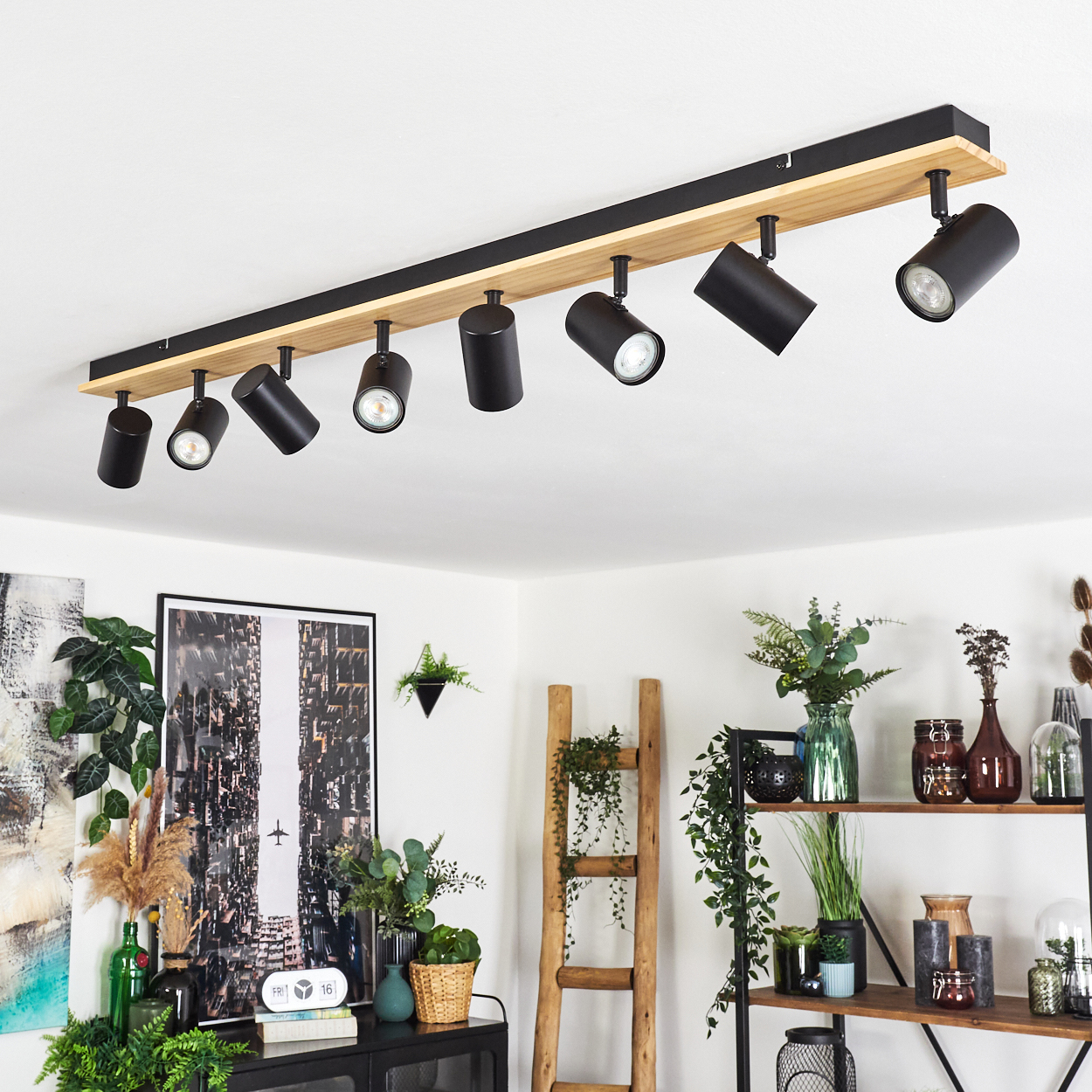 Ein modernes Zimmer mit einer Deckenleuchte. Schwarze Strahler sind an einem Holzbalken installiert. Pflanzen und eine Leiter dekorieren den Raum.