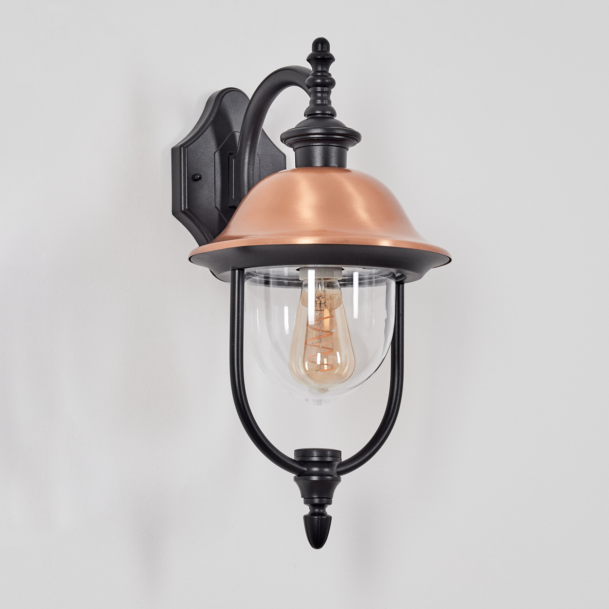 Eine Wandleuchte für den Außenbereich. Mit schwarzem Rahmen, kupferfarbenem Oberteil, Glaseinfassung und Edison-Glühbirne. An weißer Wand montiert.