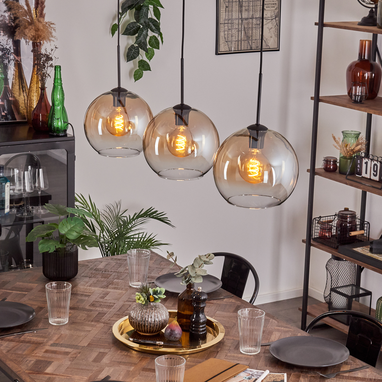 Esszimmer mit Holztisch, drei Hängelampen und Dekorationsartikeln. Enthält Gläser, Teller und Zierpflanzen.