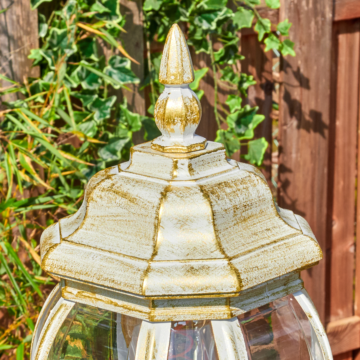 Nahaufnahme einer weiß-goldenen Außenlampe. Grünes Laub und Holzzaun im Hintergrund. Die Lampe hat eine Glaskugel.