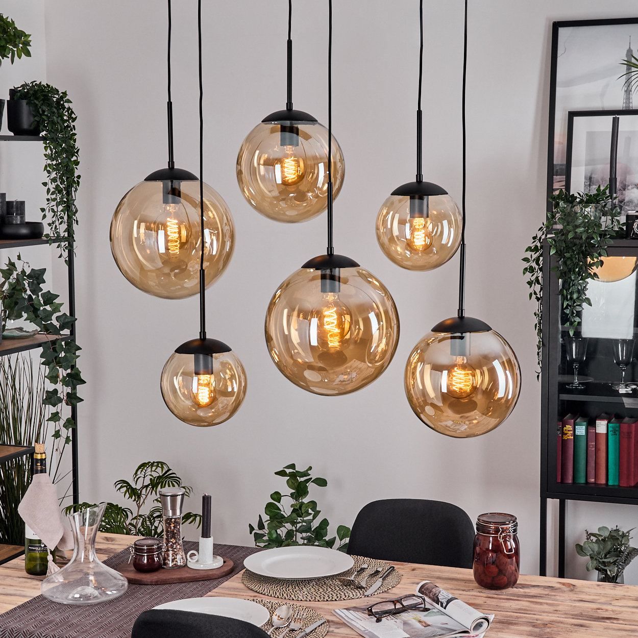 Esszimmer mit sechs bernsteinfarbenen Glaskugelleuchten. Tisch mit Tellern, Besteck und Dekorationen. Pflanzen und Regale im Hintergrund.