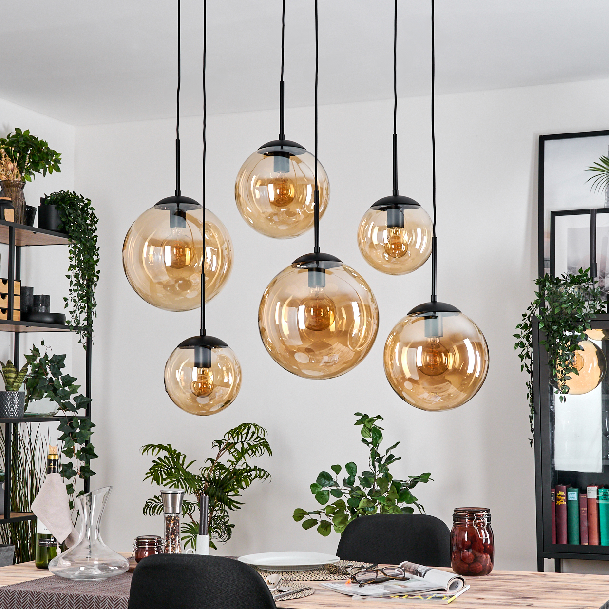 Esszimmer mit sechs hängenden, bernsteinfarbenen Glaskugelleuchten. Ein Tisch ist mit Pflanzen und Dekor gedeckt. Pflanzen und Regale sind im Hintergrund.
