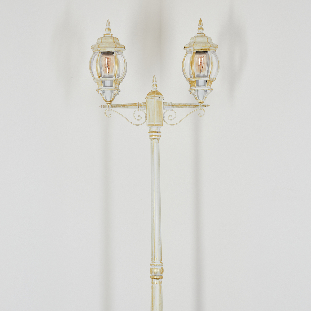 Eine verzierte, cremefarbene Straßenlaterne mit zwei glaseingeschlossenen Lichtern. Die Lampe steht vor einer weißen Wand mit Schatten.